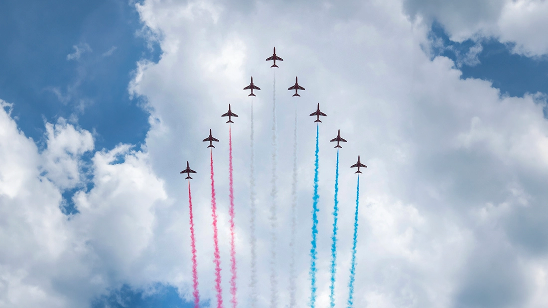 A formation of nine jet planes flying in a V-shape in the sky, emitting trails of red, white, and blue smoke against a backdrop of white clouds.