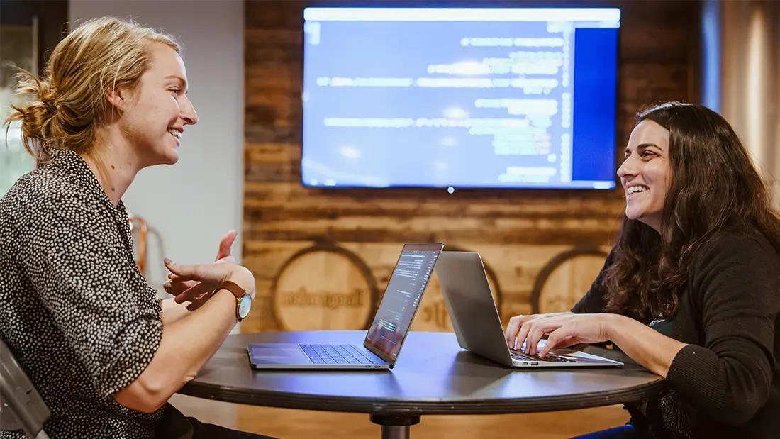 Two women collaborating on a SaaS UX project, brainstorming ideas around a laptop in a modern workspace.